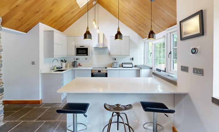 A spacious kitchen with white cabinets, slate flooring, a large island with stools, and pendant lighting beneath a vaulted wooden ceiling. A stone fea